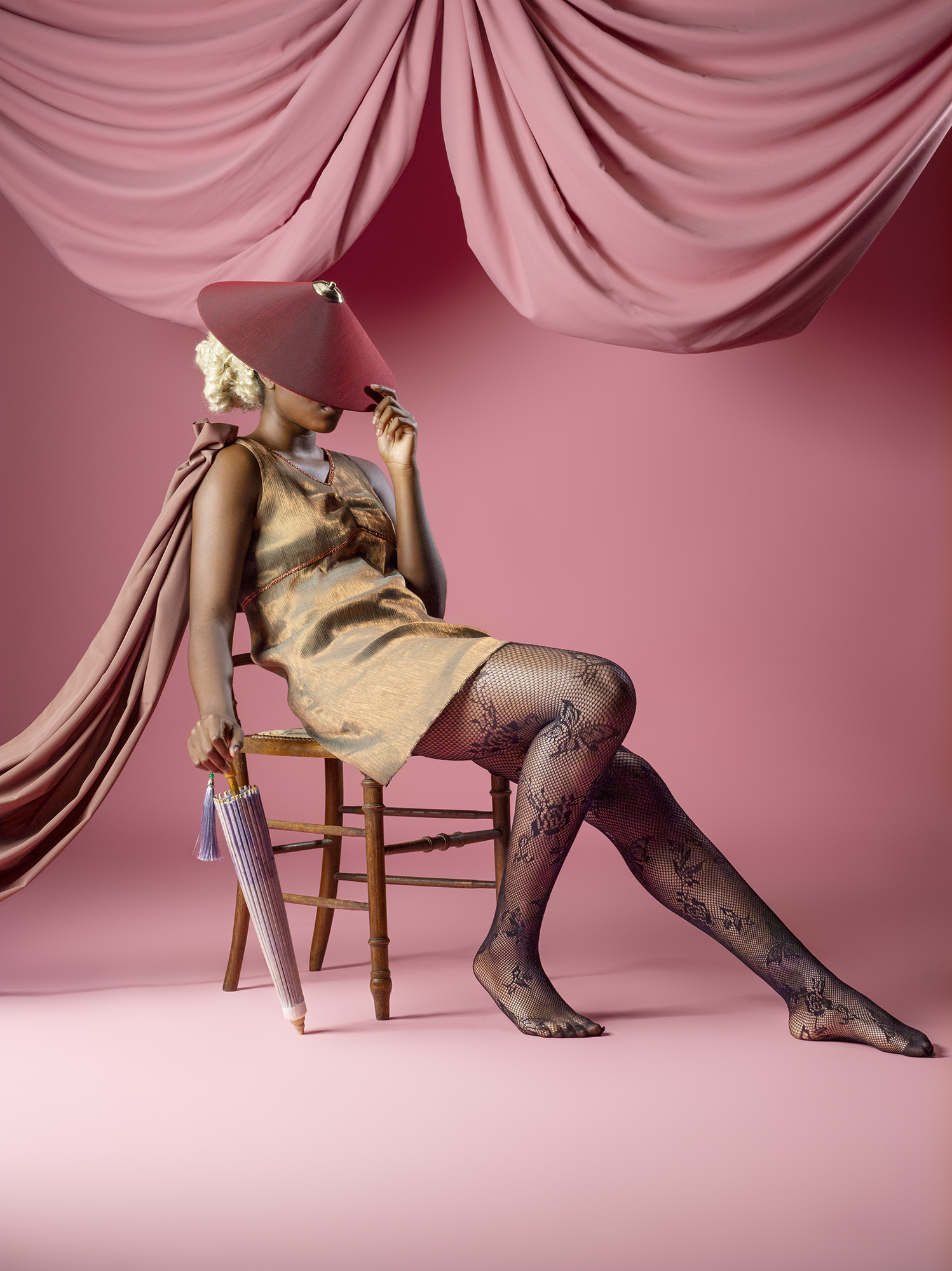 Uncanny Fashion photoshoot featuring a model on a chair wearing thrifted fashion, pink background.