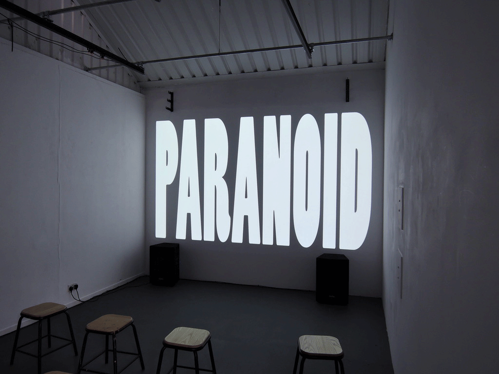 Small and dimly lit room with four stools in the foreground and the word 'Paranoid' on the wall in big letters.