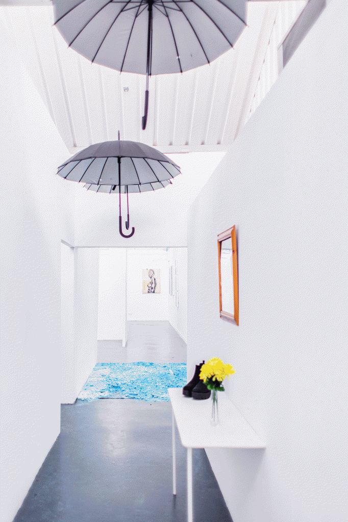 A long room with white walls umbrellas on the ceiling and a console table in the foreground. There is a small vase and a pair of shoes on the table.